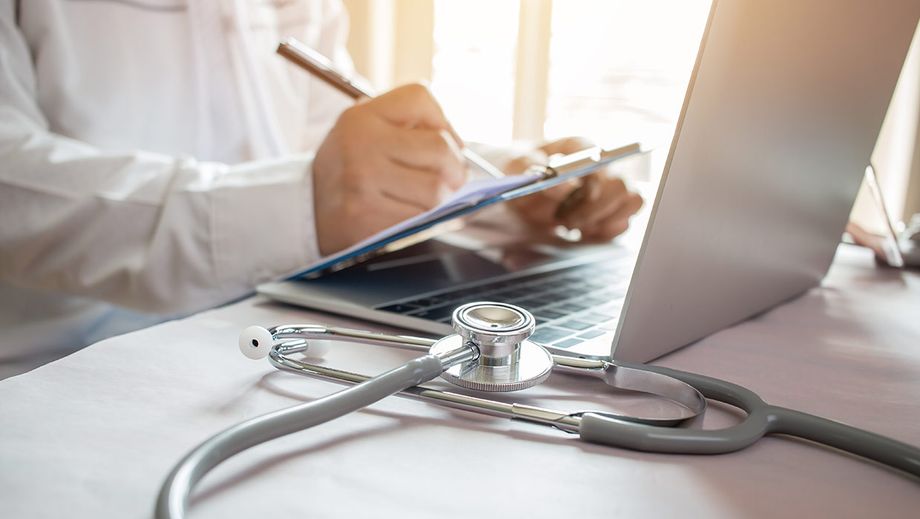Ein Arzt sitzt an einem Laptop, sein Stethoskop liegt auf dem Tisch.